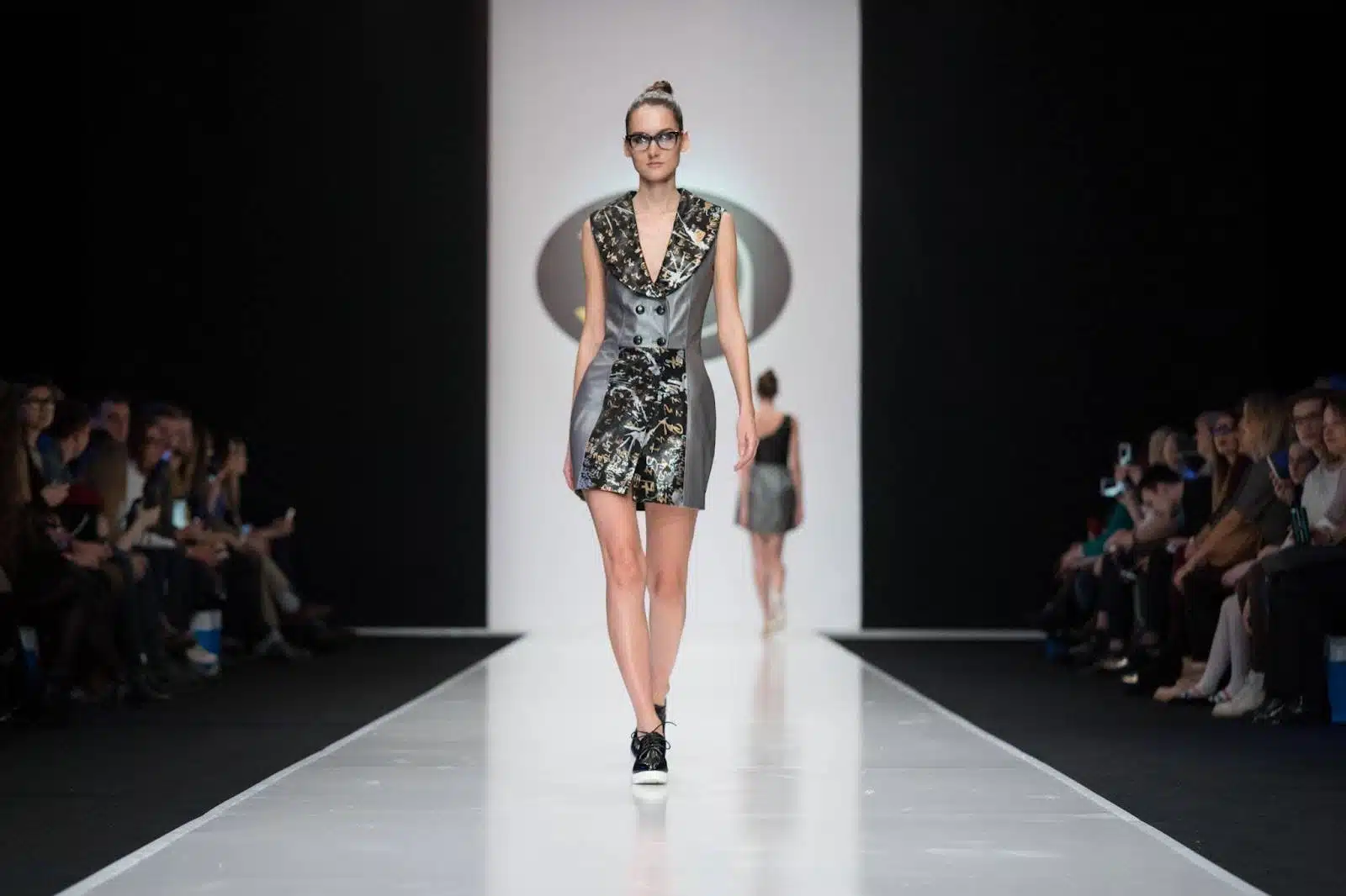 A model walks down the runway wearing a stylish, dress with glasses, as the audience watches on both sides. 