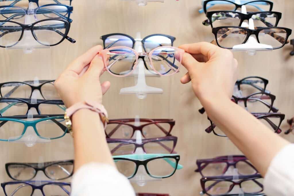 A person selects a pair of pink-rimmed glasses from a display, with various other eyeglasses in different styles and colors arranged on the wall.