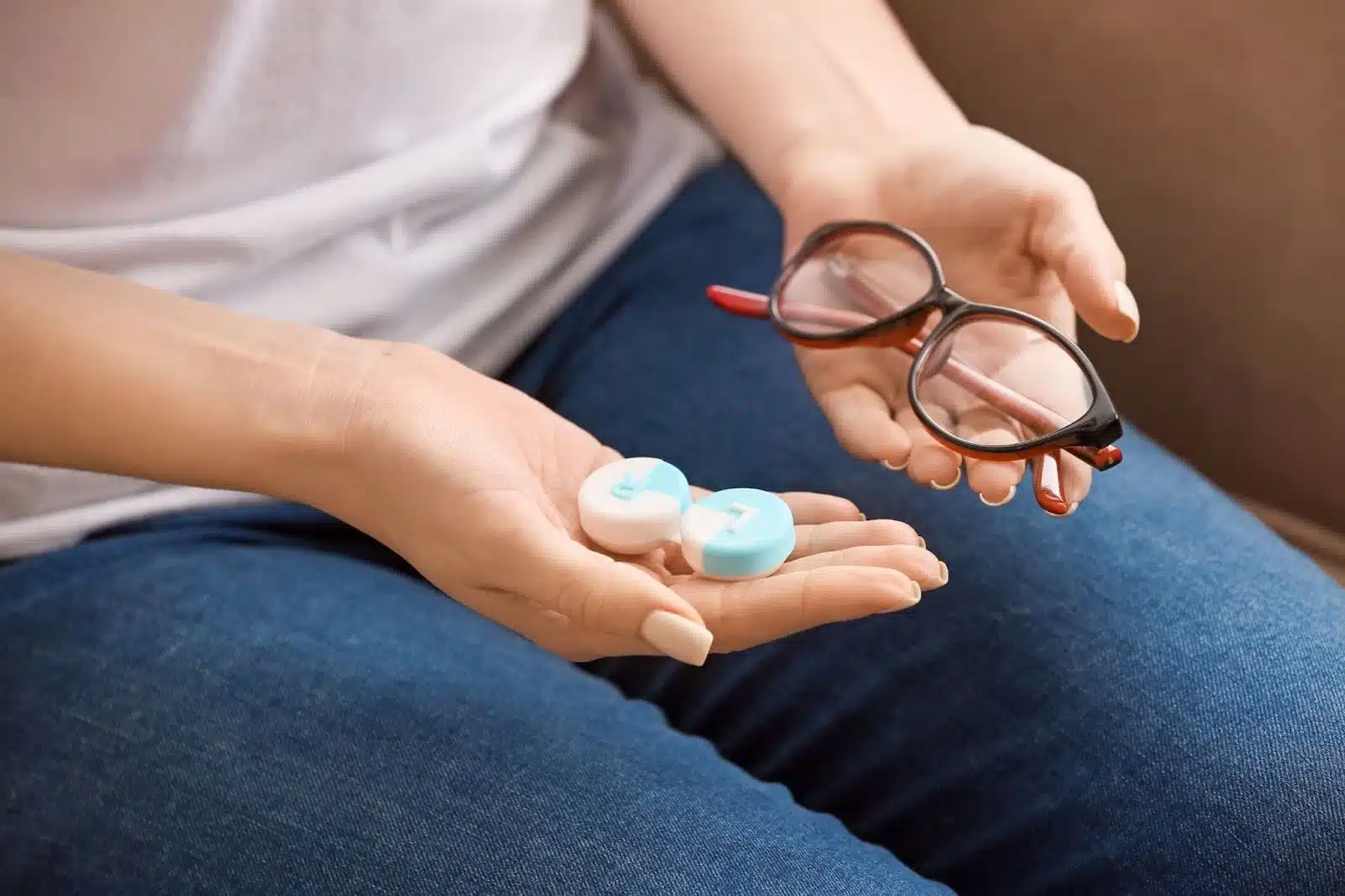 A person holds eyeglasses in one hand and a blue and white contact lens case marked "L" and "R" in the other, wearing jeans and a white shirt.