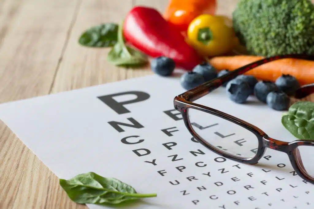 A pair of glasses rests on an eye chart, surrounded by colorful vegetables, blueberries, and spinach, emphasizing the link between diet and eye health.