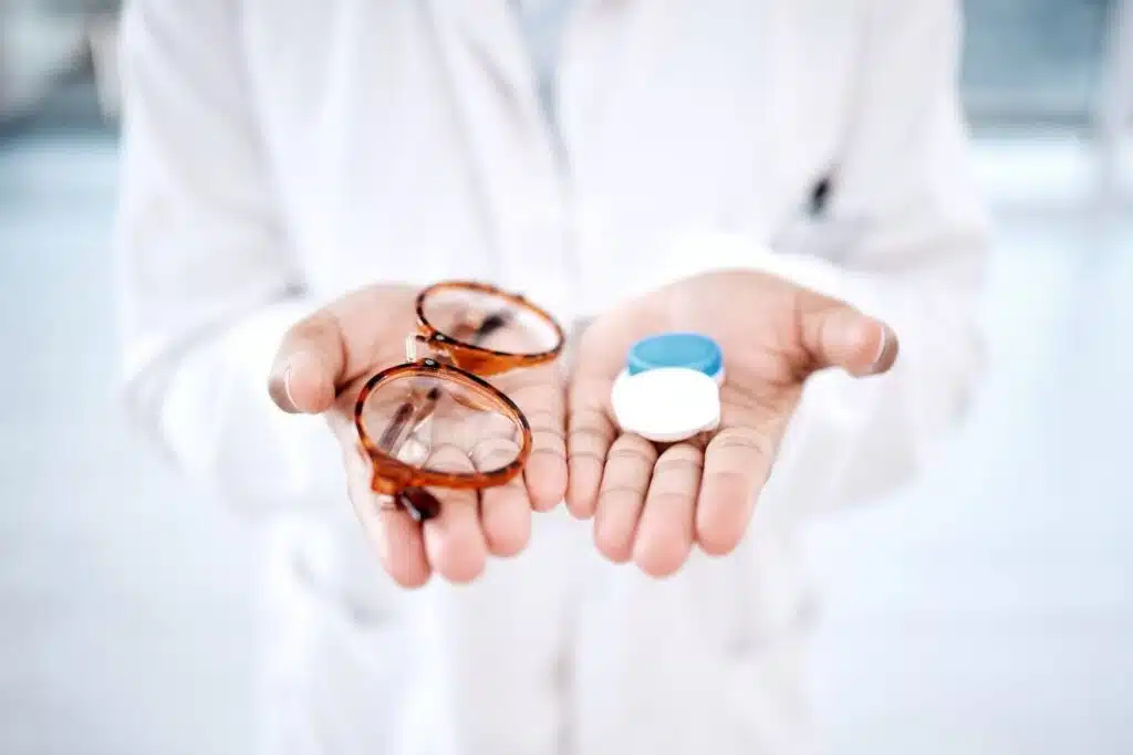 A doctor holds their hands out towards the camera, with glasses in one hand and a contacts case in the other.