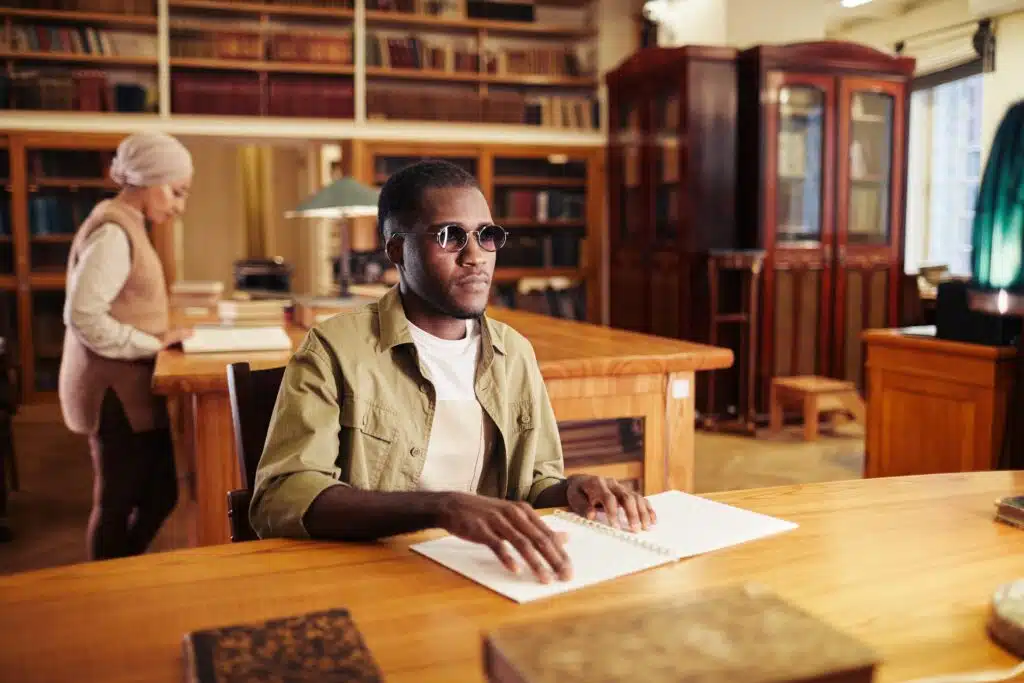 A blind, black man sits in a library reading a braille book.