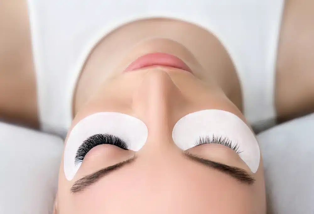 A woman lies down with eye pads placed under her eyes, showing one set of natural lashes and one set of extended lashes during an eyelash treatment.